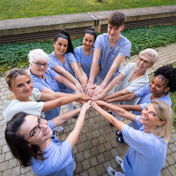 Neun Pflegepersonen legen ihre Hände zusammen und lächeln dabei in die Kamera.