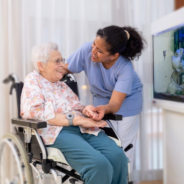 Eine Pflegefachperson und eine Bewohnerin lachen gemeinsam und halten Händchen vor einem beleuchteten Aquarium.