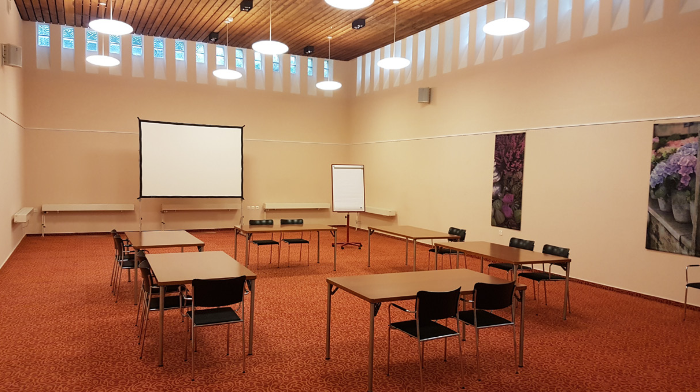 Es ist der Saal der Casavita Vincentianum zu sehen, mit rotem Teppichboden, diversen Tischen und Stühlen und hängender Deckenbeleuchtung, es sind ausserdem Flipcharts und eine Projektionsfläche zu sehen.