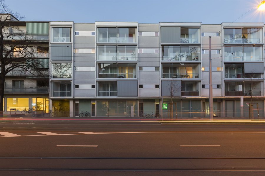Es sind die Alterswohnungen Kannenfeld zu sehen mit mehreren Etagen und Balkonen aus Glas, davor verläuft eine Strasse mit Markierungen und Tramschienen.