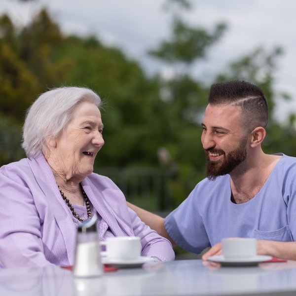 Eine Bewohnende und eine Pflegefachperson trinken lächelnd gemeinsam einen Kaffee im Grünen.