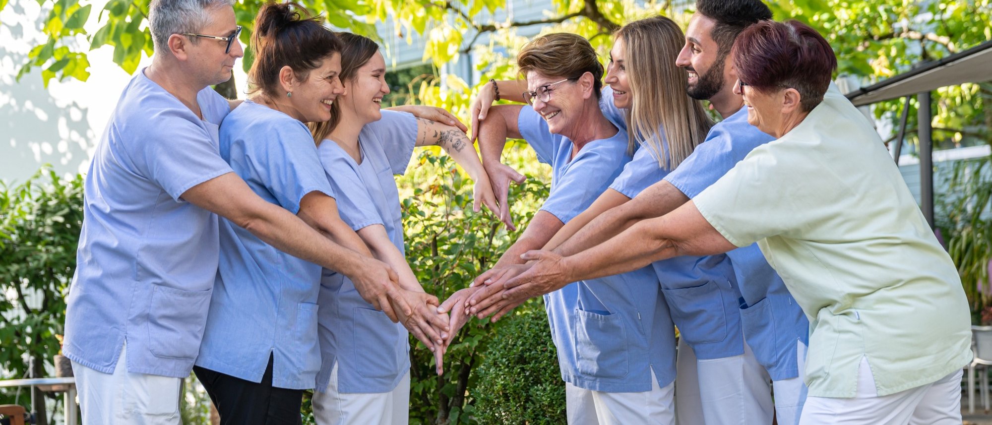 Sieben Pflegepersonen formen mit ihren Armen gemeinsam ein grosses Herz und lächeln sich dabei gegenseitig an.
