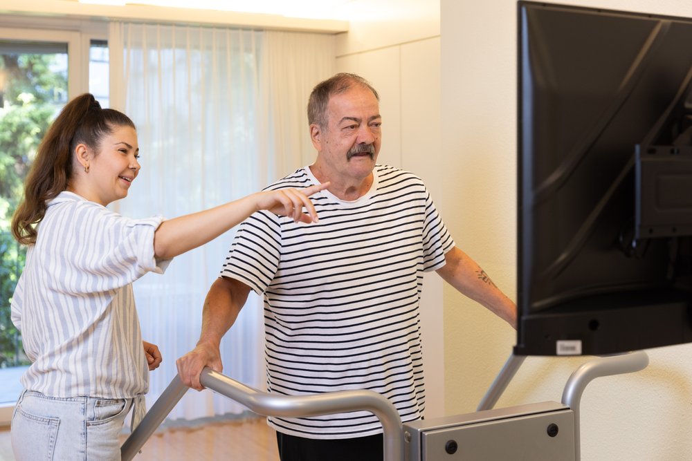 Es sind ein Bewohnender und eine Pflegende zu sehen, der Bewohnende steht auf dem Gleichgewichtstrainingsgerät «Dividat Senso» und die Pflegende zeigt mit dem Zeigefinger auf den Bildschirm des Geräts.