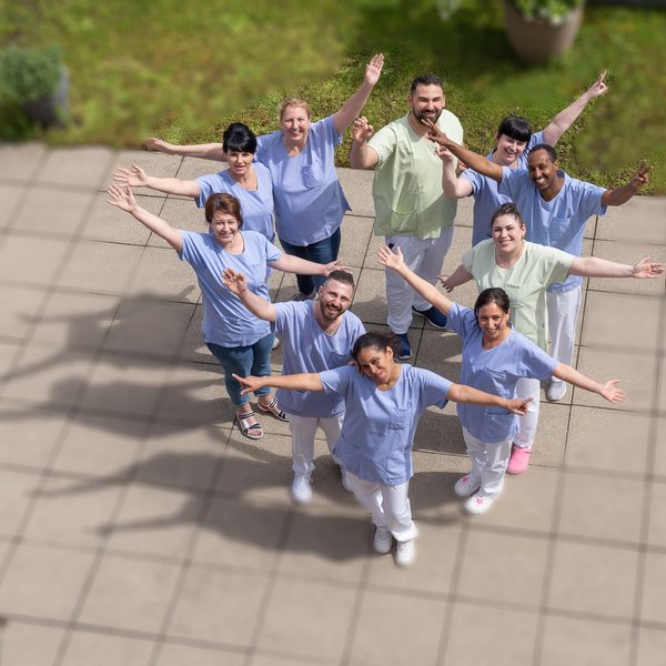 Es sind diverse Pflegepersonen zu sehen welche lächelnd in die Kamera ihre Arme ausstrecken.