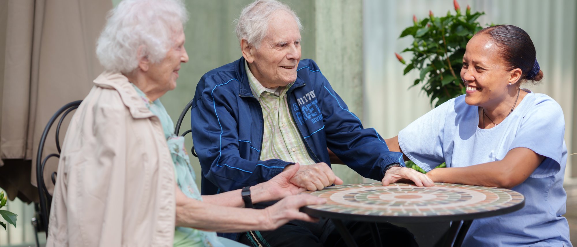 Ein Bewohnendenpaar welches Händchen hält, sitzt zusammen an einem Tisch mit einer Pflegeperson welche lächelnd ihren Arm auf den Rücken des Bewohnenden hält.