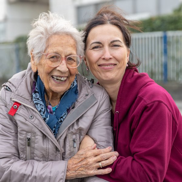 Lächelnde Bewohnende und Mitarbeitende umarmen sich vertraut, die Bewohnende hält dabei wertschätzend die Hand der Mitarbeitenden.