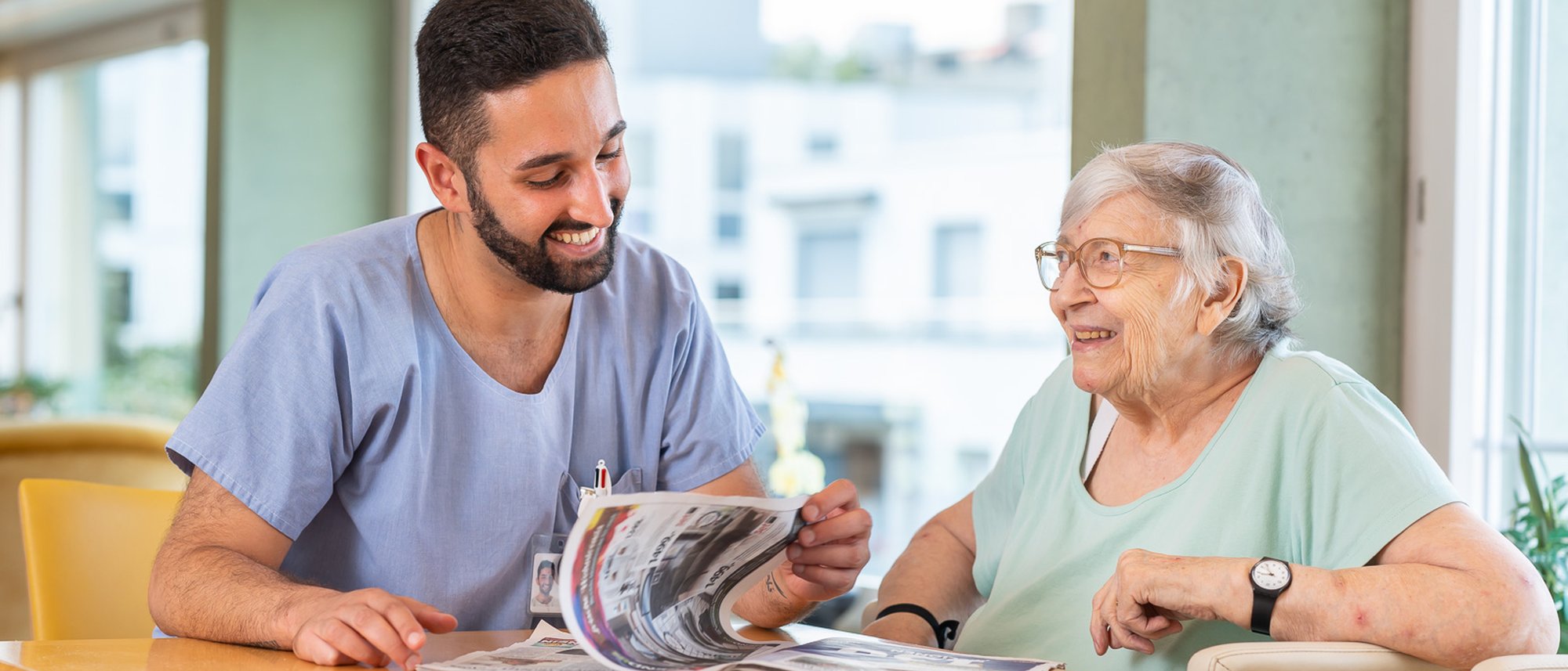 Eine Pflegekraft und eine Bewohnerin sitzen an einem Tisch und schauen sich eine Ausgabe des Casavita Magazins an. Im Hintergrund ist ein Fenster zu erkennen.