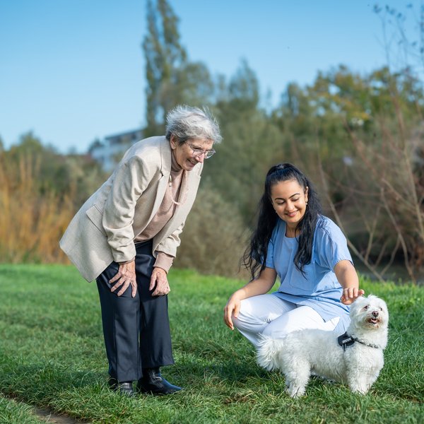 Eine Bewohnerin und eine Pflegekraft sind bei blauem Himmel unten an der Birss. Die Pflegekraft kniet im Rasen und streichelt einen kleinen weissen Hund.