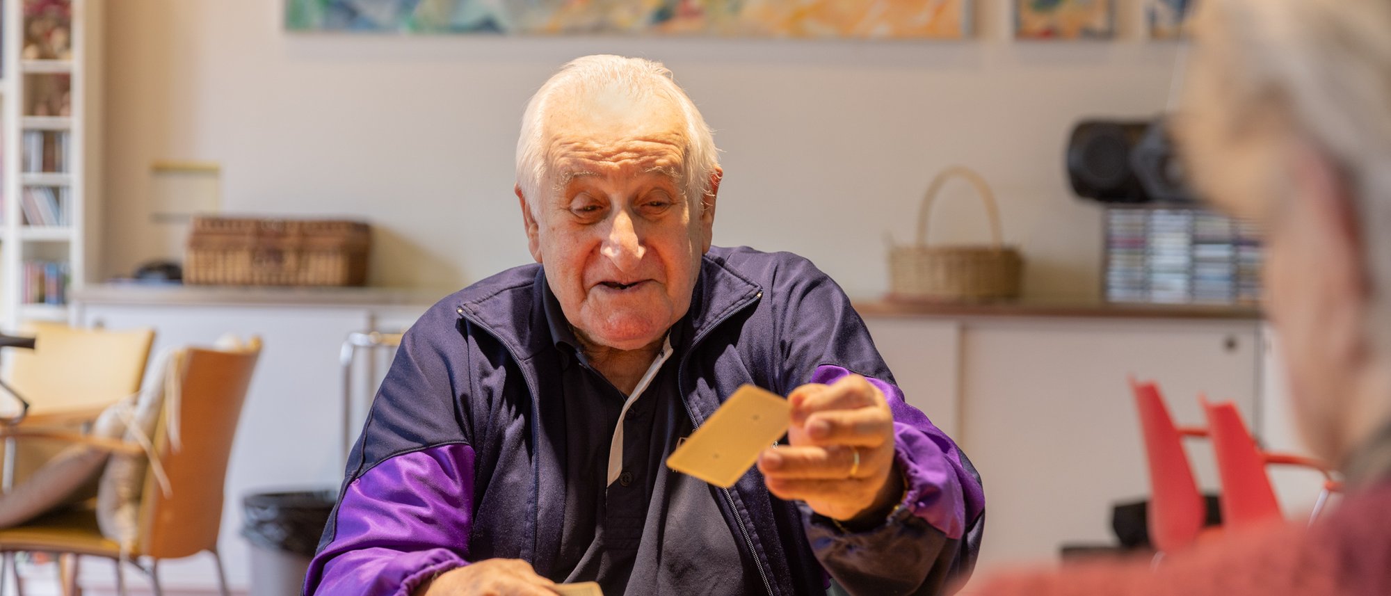 Ein Bewohnender mit einer lila Sportjacke spielt Karten mit einer Bewohnenden welche aber nur von hinten zu sehen ist.
