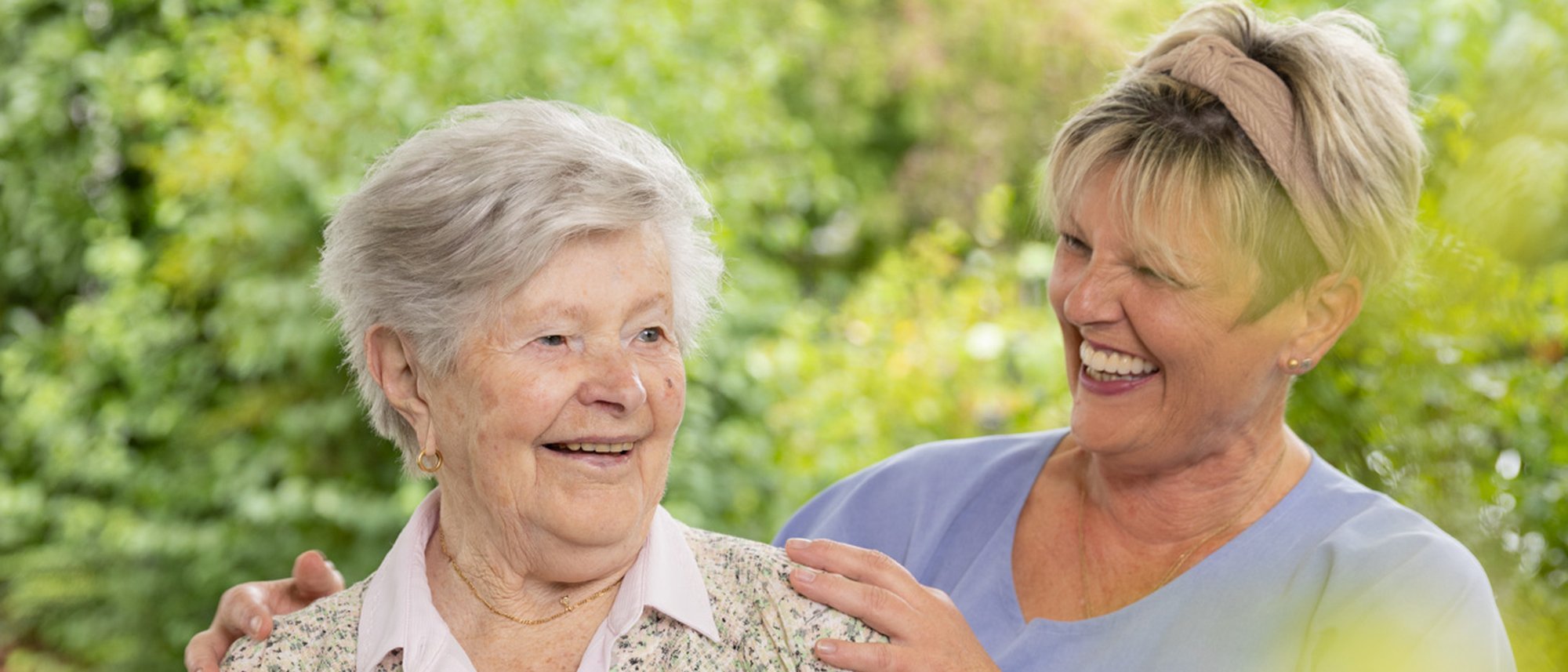 Eine Pflegende hat ihre Hände auf die Schultern einer Bewohnenden gelegt und lächelt sie dabei freundlich an. Sie befinden sich in einem Garten.
