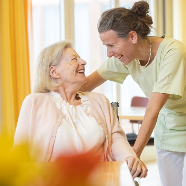 Eine Pflegeperson stützt sich mit einem Arm auf den Rollstuhl einer Bewohnenden und sie lachen sich gegenseitig an.