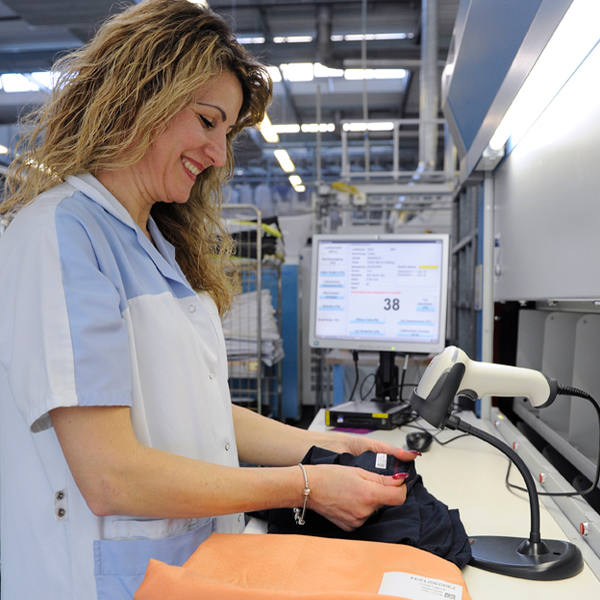 Eine Mitarbeitende in blauer Arbeitskleidung scannt Wäschestücke mit einem Scanner an einem Arbeitsplatz mit einem Computer, im Hintergrund sieht man einen Metal-Wäschewagen.