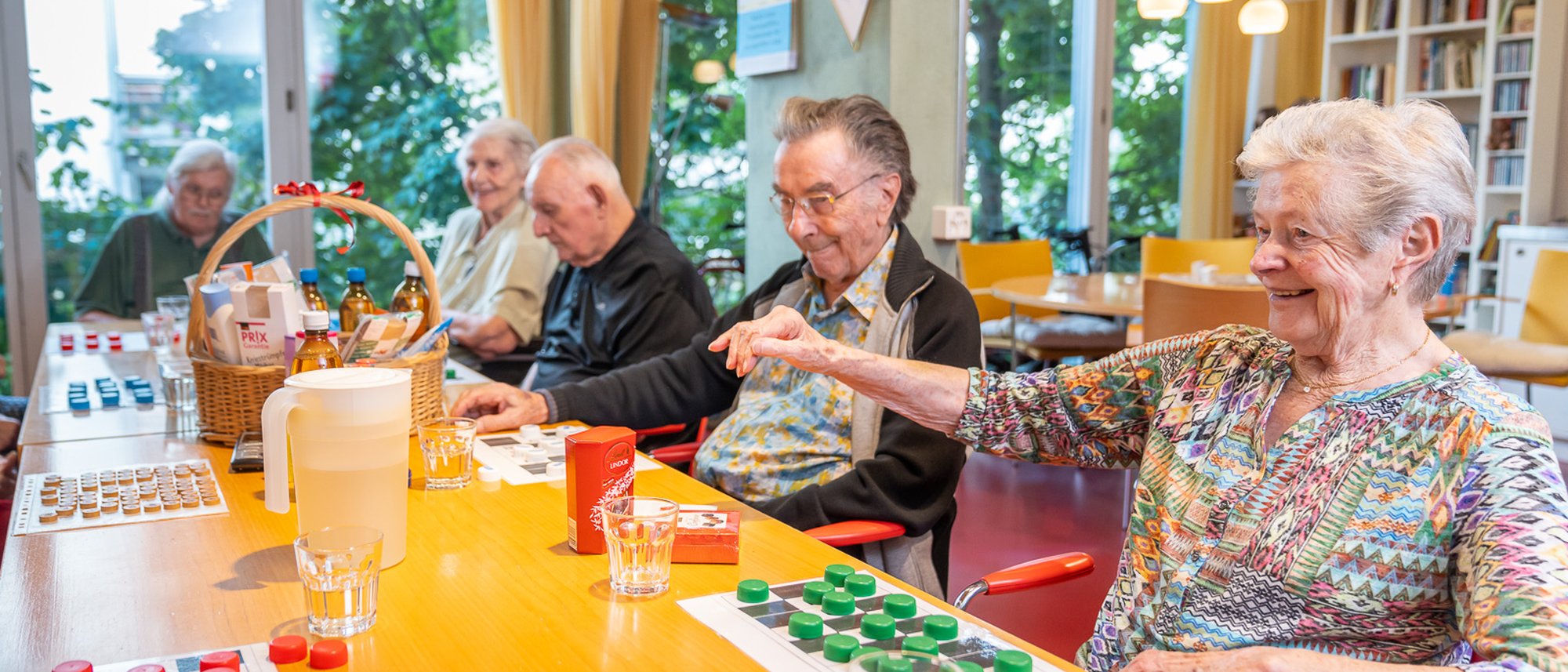 Eine Gruppe von Bewohnenden spielt gemeinsam Lotto an einem langen Tisch. In einem Korb in der Mitte sind verschiedene Preise zu sehen.