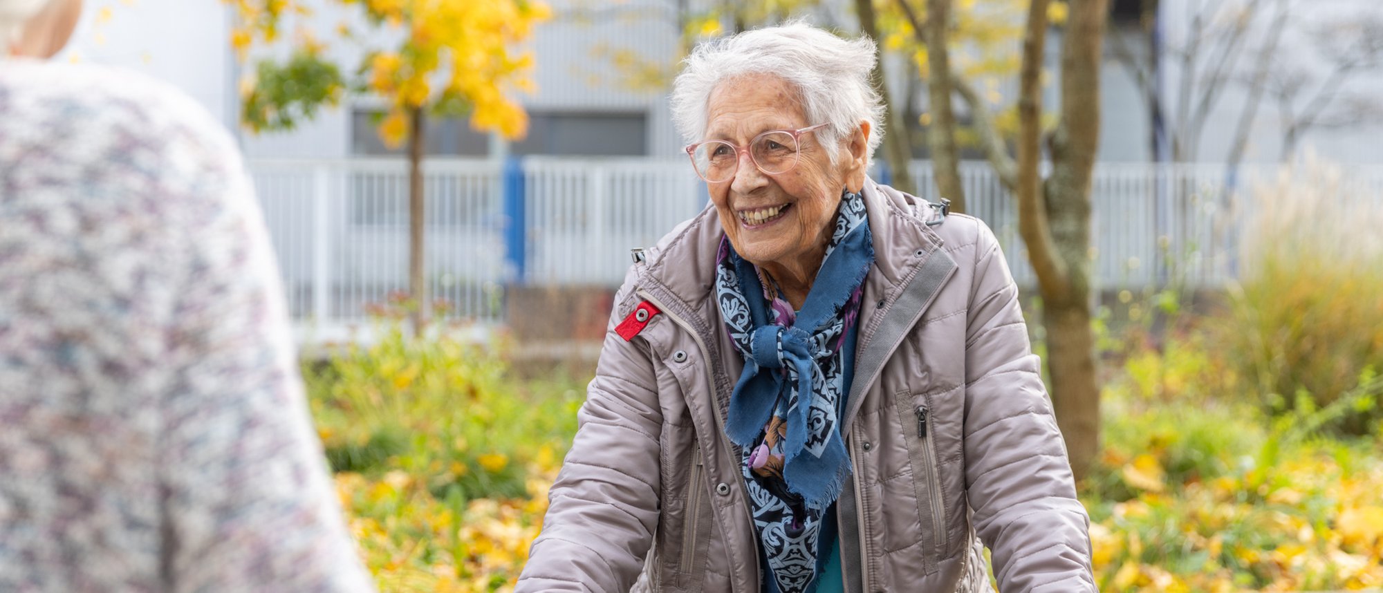 Es ist eine lächelnde Bewohnende zu sehen welche ihren Rollator festhält, im Hintergrund ist eine herbstliche Parklandschaft erkennbar.