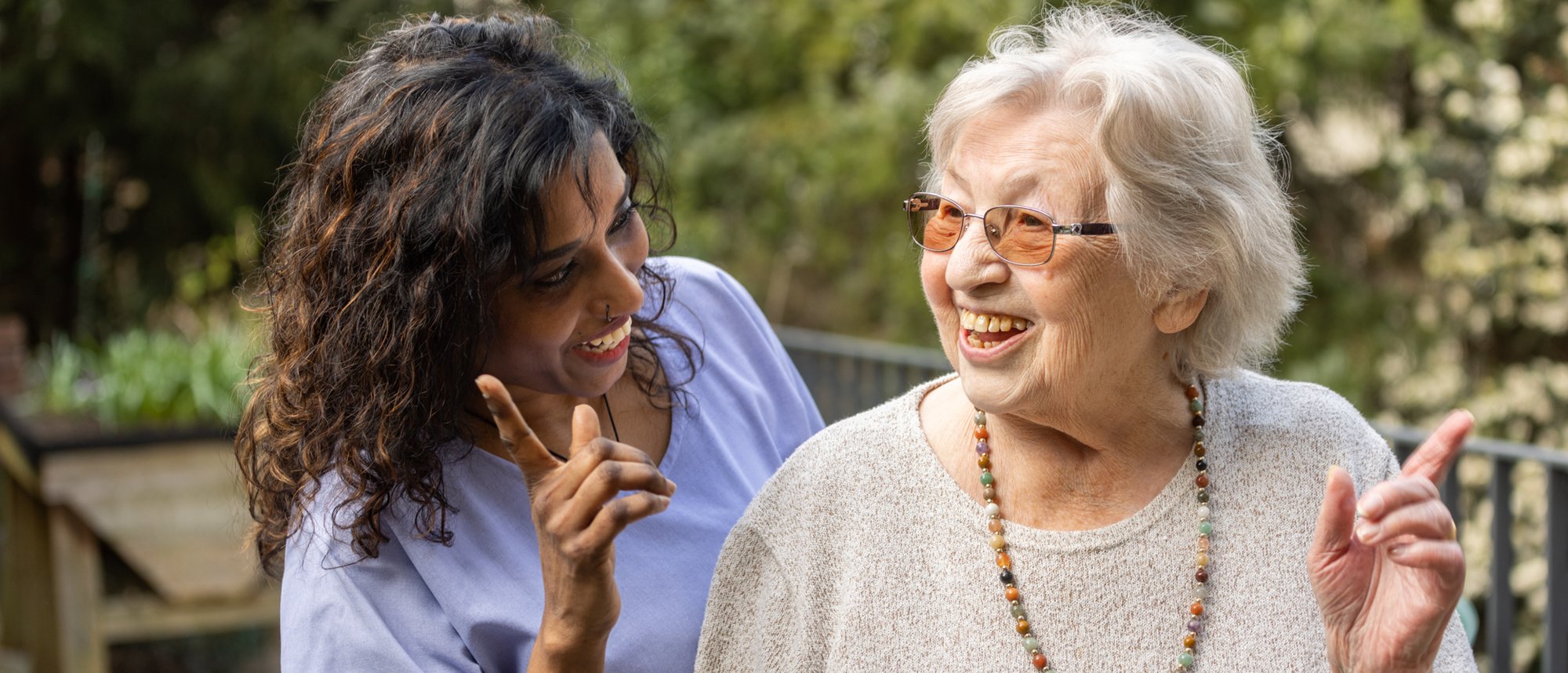 Eine Pflegeperson und eine Bewohnende stehen im Freien nebeneinander und zeigen jeweils mit dem Finger in eine Richtung.