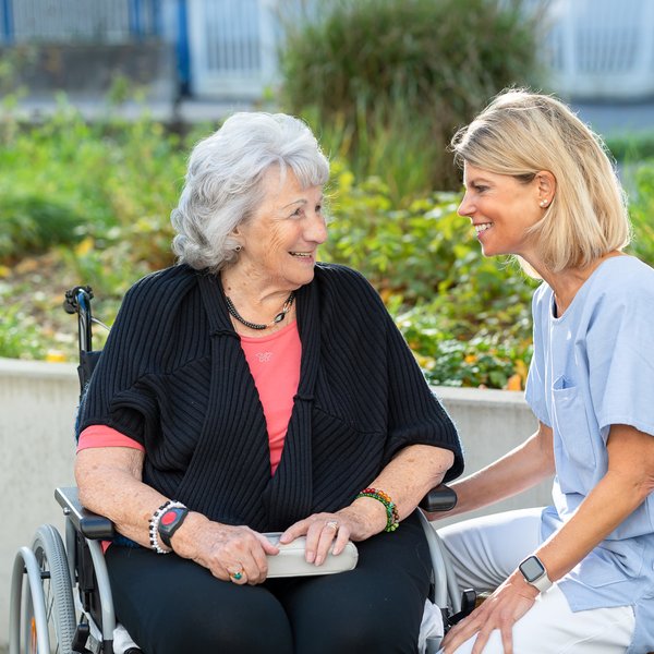 Eine Pflegeperson und eine Bewohnende im Rollstuhl sitzen gemeinsam im Aussenbereich der Casavita Lehenmatt und lächeln, im Hintergrund sind grüne Pflanzen zu sehen.