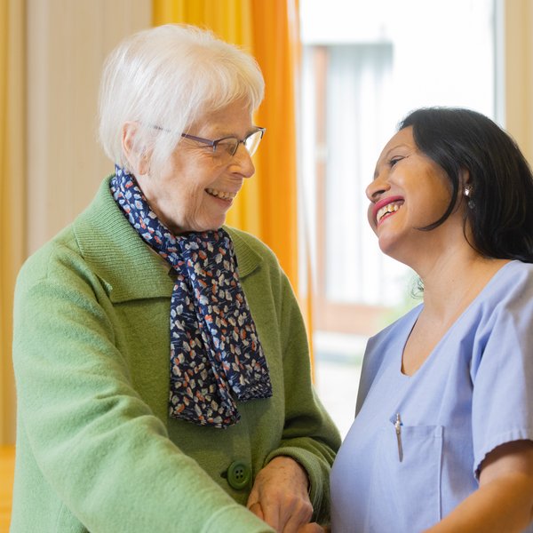 Eine Pflegeperson und eine Bewohnende lächeln sich an, im Hintergrund ist ein grosses Fenster und gelbe und orange Vorhänge zu sehen.