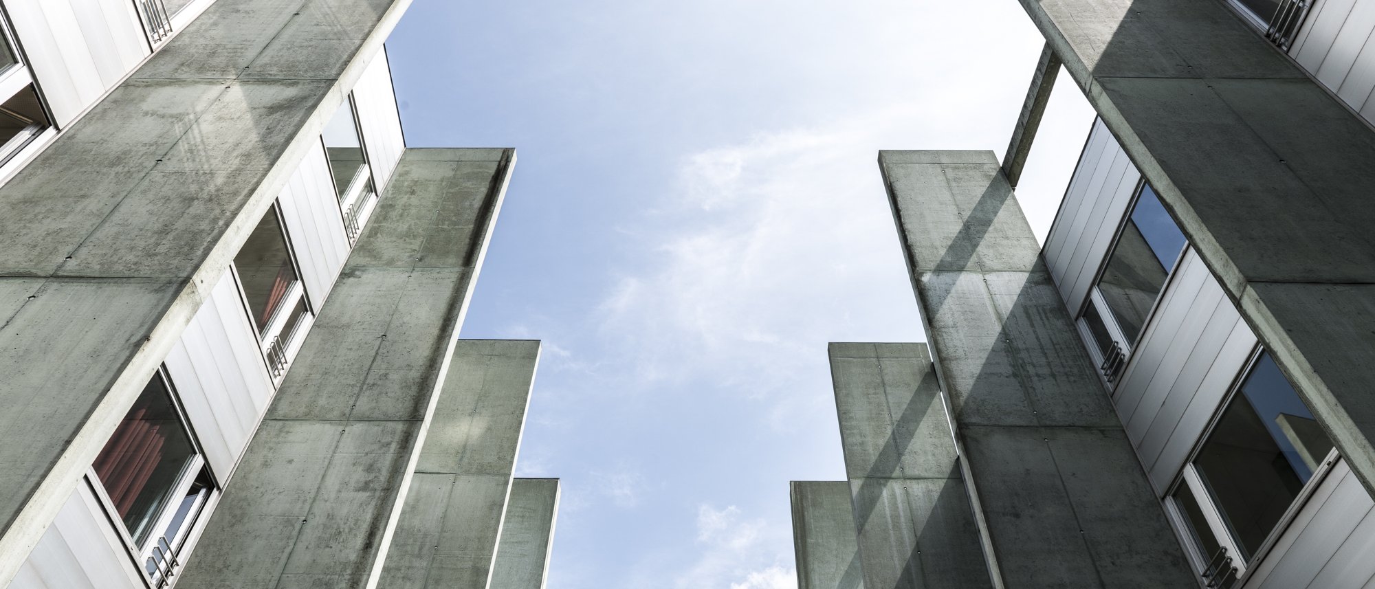 Blick nach oben zwischen zwei modernen Gebäudeflügeln aus Beton und Glas, mit Fenstern und freiem Himmel in der Mitte.