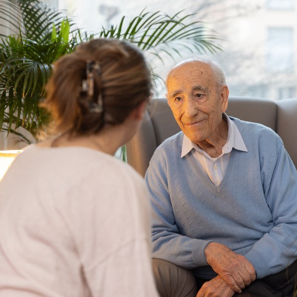 Eine Mitarbeitende ist in einem Gespräch mit einem Bewohnenden, sie sitzen in einem Zimmer mit grossen Fenstern, umgeben von Grünpflanzen.