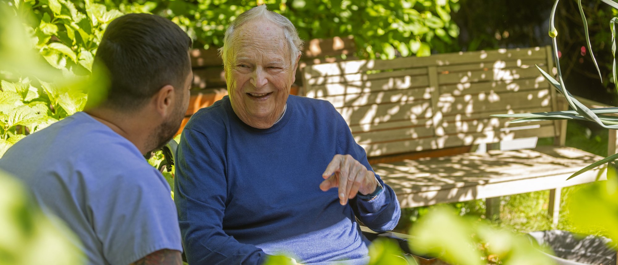 Ein Bewohner sitzt im Garten der Casavita Hasenbrunnen und ist im Gespräch mit einer Person aus der Pflege verwickelt. Dabei hebt er gestikulierend seine Hand.