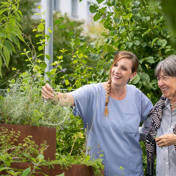 Eine Pflegeperson zeigt einer Bewohnenden eine Rosmarinpflanze im Topf während die Bewohnende einen Arm um die Pflegeperson hat und ein kleines Sträuchchen davon in der Hand hält.