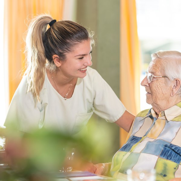 Pflegeperson spricht mit einer Bewohnenden an einem Esstisch im Innenbereich, während sie sich die Hand halten.