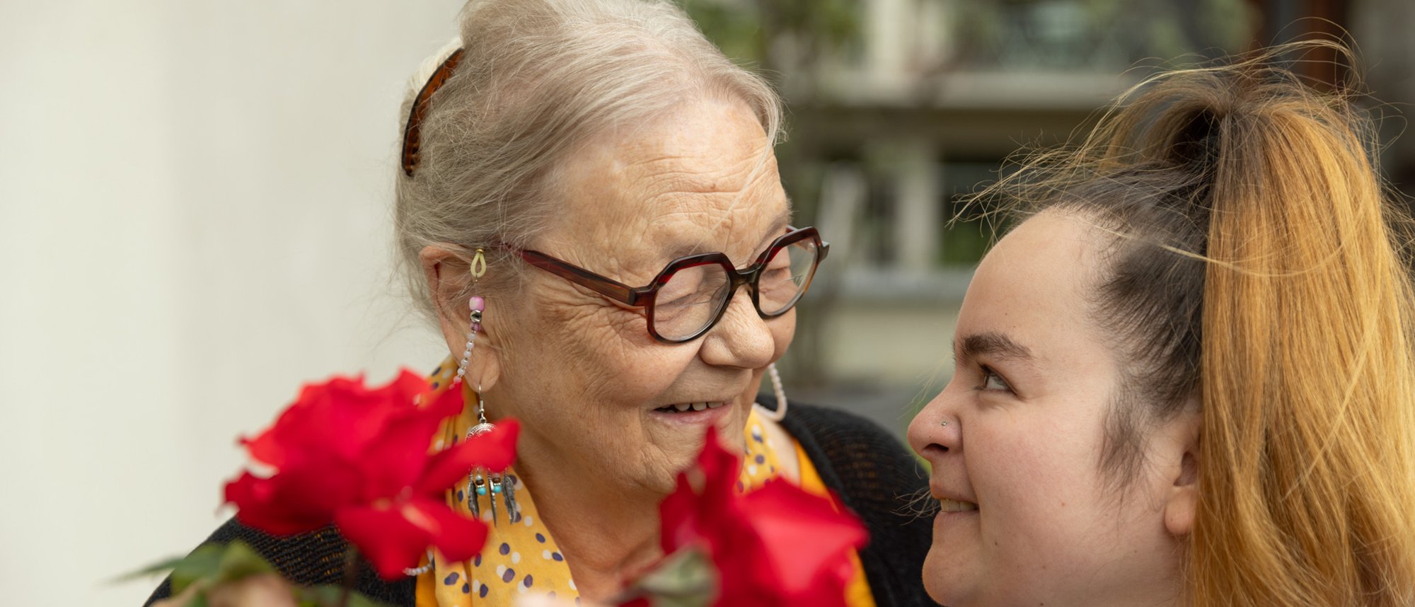 Im Vordergrund ist eine rote Rose zu erkennen und dahinter eine Bewohnende, welche sich lächelnd einer Pflegeperson zugewendet hat.